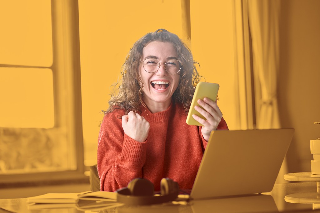 A woman with curly hair and glasses smiles joyfully while holding a smartphone, sitting at a desk with a laptop and headphones in a bright room.